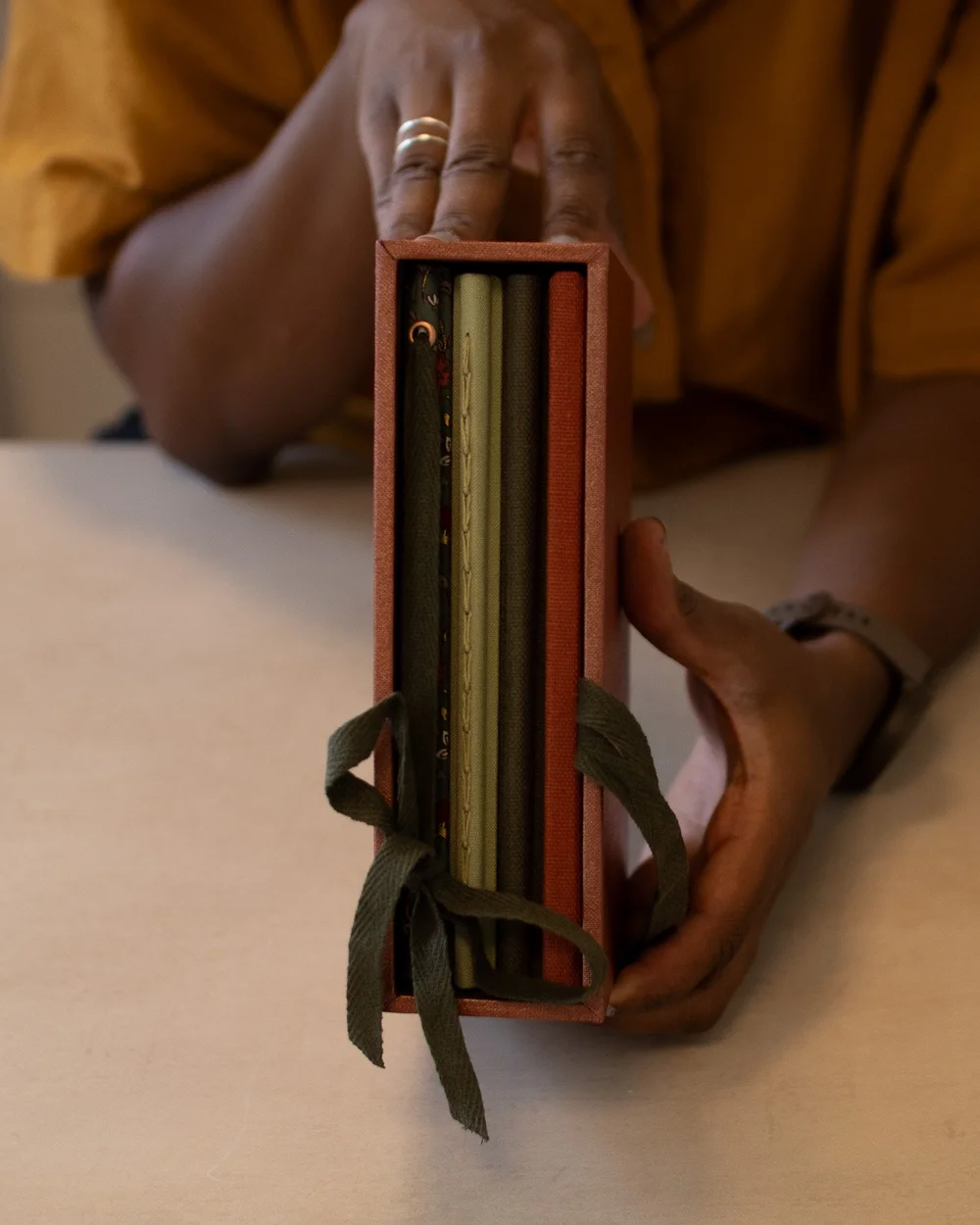 Hands holding a stack of bound books tied with cord, resting in an open rust-orange box